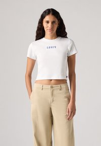 Balta trumpa marškinėliai su trumpomis rankovėmis, centre yra mėlynas "LEVI'S" užrašas. Sujungti su šviesiai smėlinėmis plačiomis kelnėmis.