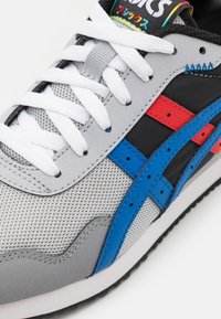 Grå sportskor med mesh-textur, vita snören och färgade accenter i rött och blått. Påtaglig logotyp på tungan och sidan.