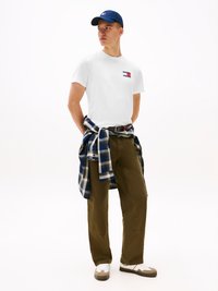 White t-shirt with logo, plaid flannel shirt tied at waist, olive green pants, blue cap, and white sneakers with brown accents.