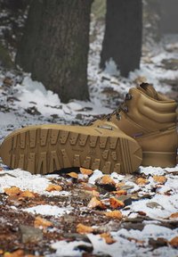 Bruine wandelschoenen met robuuste zolen die rusten op een besneeuwde bosbodem bedekt met gevallen bladeren en stenen nabij boomstammen.