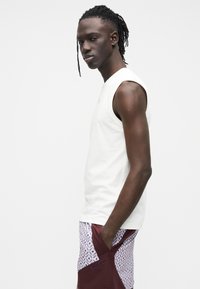 Chemise blanche sans manches avec un col rond, associée à un short bordeaux avec un motif géométrique et des panneaux latéraux contrastants.