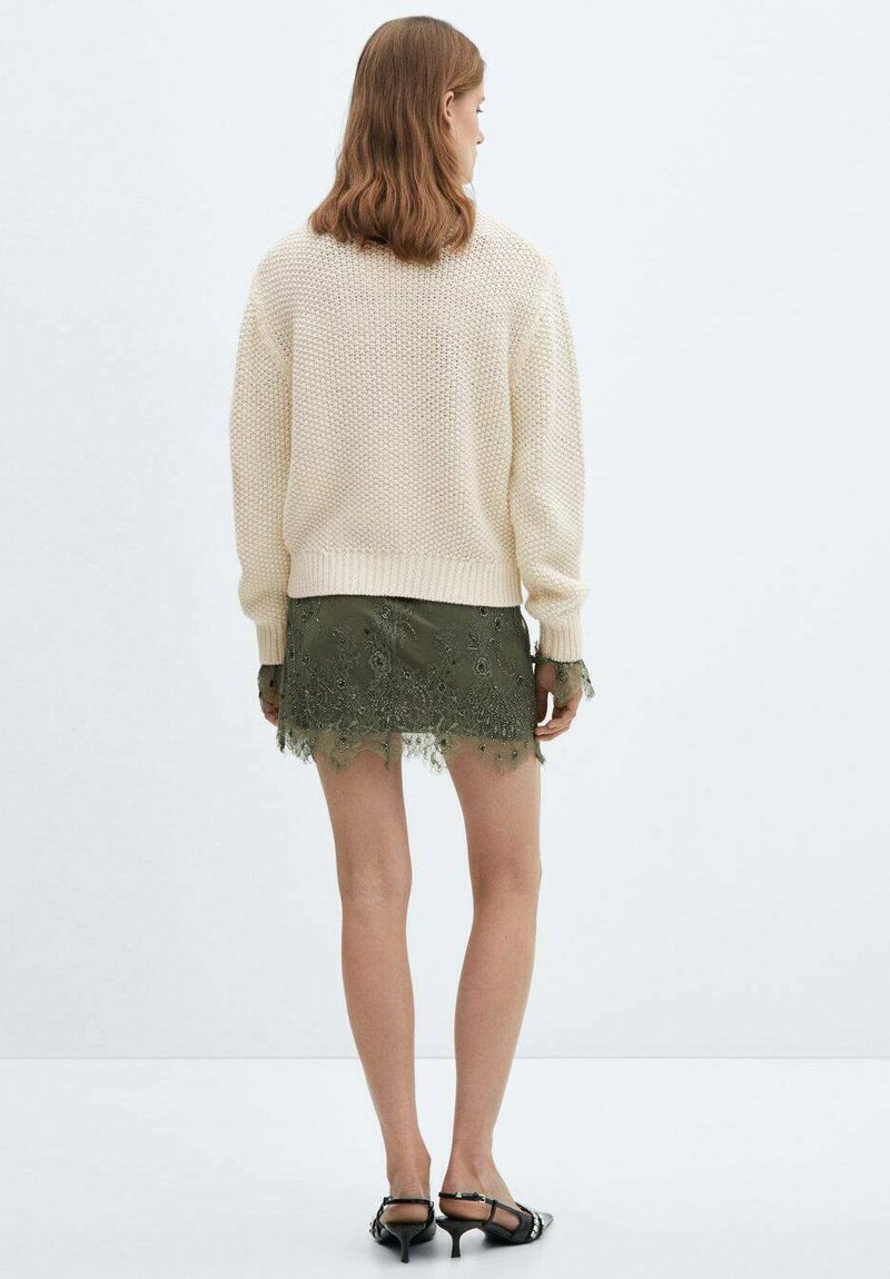 Pull en tricot crème avec une texture épaisse, associé à une mini-jupe verte brodée avec des détails en dentelle et des sandales à lanières noires.