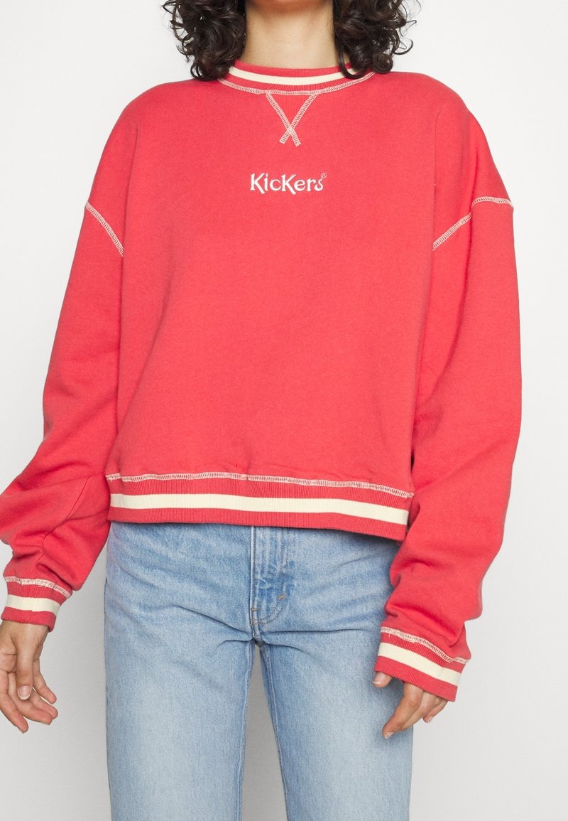 Personne portant un sweat Kickers rouge avec des poignets et un ourlet rayés blancs, associé à un jean taille haute bleu clair.