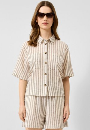 Frau trägt braune Cat-Eye-Sonnenbrille und beige gestreiftes kurzärmeliges Hemd mit passenden Shorts.