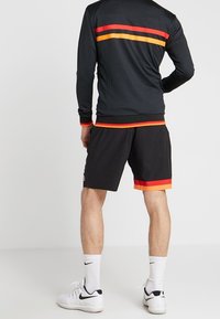 Man som bär en svart sportjacka och shorts med röda och oranga ränder, vita Nike-strumpor och vita sneakers, står med ryggen mot kameran.