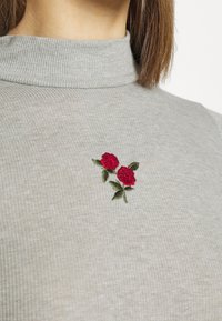 Grå ribbad turtleneck överdel med en liten broderad design av två röda rosor med gröna blad på vänster sida av bröstet.