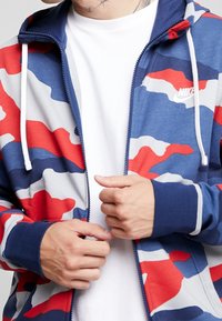 Röd, blå och grå camouflage zip-up hoodie med en dragsko på huvan, ribbade ärmar och en vit Nike-logotyp på vänster bröst. Material i bomullsblandning.