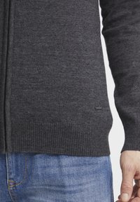 Cardigan en tricot gris foncé avec un ourlet et des poignets côtelés, doté d'une texture lisse et d'un petit logo en bas. Porté sur un jean bleu.