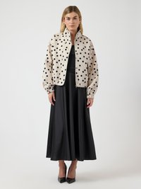 Γυναίκα που φοράει μπεζ καπιτονέ μπουφάν με πουά, μαύρο αμάνικο τοπ, μαύρη μίντι φούστα και μαύρες μυτερές ψηλοτάκουνες γόβες, στέκεται μπροστά σε λευκό φόντο.