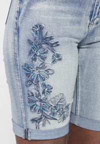 Shorts en denim bleu clair avec des revers, présentant une broderie florale complexe en bleu foncé et turquoise d'un côté.