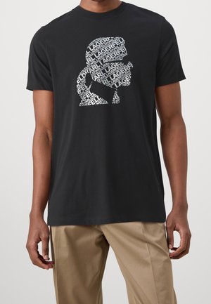 Mężczyzna w czarnym t-shircie z białą sylwetką logo Karla Lagerfelda, wypełnioną powtarzającym się tekstem z nazwą marki, zestawionym z beżowymi spodniami.