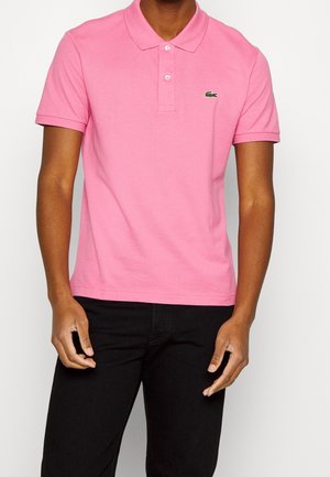 Man in een roze polo met korte mouwen en een klein groen krokodillenlogo op de linkerborst, gecombineerd met zwarte broek.