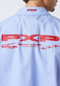 Homme portant une chemise à fines rayures bleues et blanches avec un grand logo rouge "PXP" et "Project X Paris" imprimé au dos.