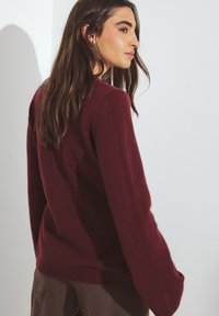 Burgundy stickad tröja, långa ärmar, ribbad nederkant, slät textur, avslappnad passform, bärs med bruna byxor mot en ljus bakgrund.