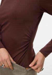 Brun langermet topp med rynket detalj på siden, laget av et glatt stoff. Delvis stukket inn i grå midjebånd med glidelås.