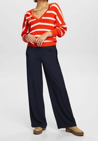 Rot-weißer gestreifter Pullover mit V-Ausschnitt, kombiniert mit navyblauen weiten Hosen. Weicher Stoff, entspannte Passform und lässiger Stil. Braune Knöchelstiefel.