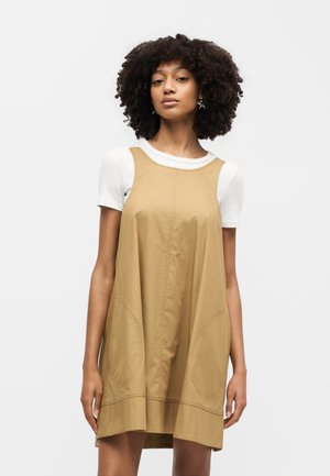 Γυναίκα με σγουρά μαλλιά που φοράει καφέ αμάνικο φόρεμα πάνω από λευκό κοντομάνικο πουκάμισο, στέκεται μπροστά σε απλό φωτεινό φόντο.