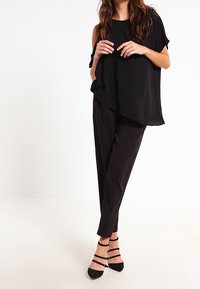 Top nero senza maniche con vestibilità ampia, abbinato a pantaloni neri su misura. Tacchi neri con cinturino alla caviglia completano l'outfit. Tessuto con texture liscia.