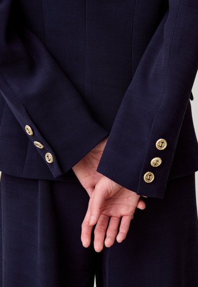 Abito sartoriale blu navy con tessuto strutturato; presenta bottoni dorati a forma di ancora sui polsini, mani intrecciate dietro la schiena.