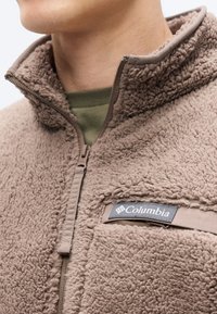 Καφέ μπουφάν από φλις με μισό φερμουάρ και υφή στην επιφάνεια· διαθέτει φερμουάρ και μαύρο έμβλημα Columbia στο στήθος.