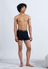 Tânăr stând desculț, îmbrăcat în boxeri negri Calvin Klein, pe un fundal simplu deschis.