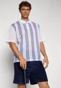 Hvit mesh polo-skjorte med blå vertikale striper, en halv glidelås-kraav og en liten logo, sammen med marineblå kordfløyelsshorts.