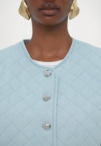 Jasnoniebieska pikowana kurtka z okrągłym kołnierzem i srebrnymi napami, o miękkiej teksturze materiału i diamentowym wzorze.