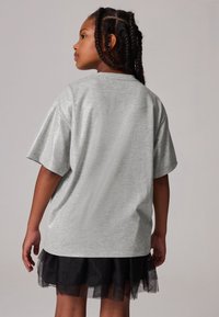 Světle šedé oversized tričko s krátkým rukávem a hladkou strukturou, v kombinaci s černou tylovou sukní. Pohled zezadu ukazuje uvolněný střih trička.