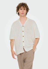 Knit button-up shirt in cream with short sleeves, featuring a v-neck, perforated texture, and brown buttons along the front. Trousers in light brown.