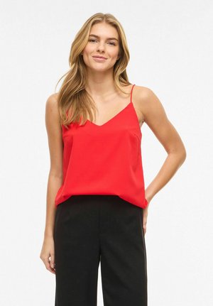 Giovane donna con lunghi capelli biondi, indossa un top rosso senza maniche e pantaloni neri, in piedi davanti a uno sfondo bianco semplice.