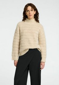 Ljusbeige stickad tröja med hög krage och randig struktur, i kombination med svarta byxor med ett asymmetriskt mönster och sidoknapp.