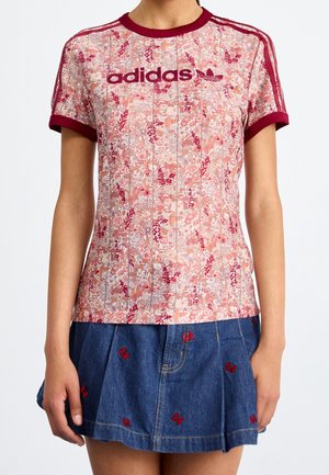 Femme portant un t-shirt Adidas rose à motifs floraux avec une bordure bordeaux et trois bandes, associé à une jupe en jean ornée d'une petite broderie de papillons rouges.