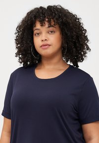 Curly-haired person with hoop earrings wearing a navy blue t-shirt, looking directly at the camera against a light background.