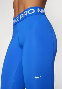 Niebieskie legginsy sportowe o dopasowanym kroju, gładkiej fakturze, elastycznym pasku w talii z napisem "NIKE PRO" w białym kolorze oraz małym logo przy kostce.