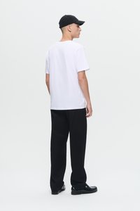 Junger Mann mit einem weißen T-Shirt, schwarzen Hosen, schwarzen Schuhen und einer schwarzen Mütze, steht und schaut von hinten auf einem einfarbigen hellen Hintergrund.