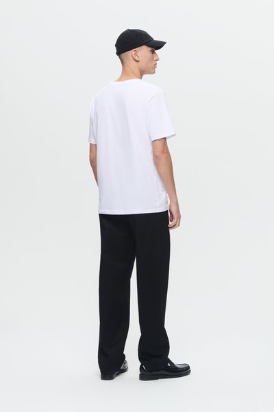 Jonge man in een wit T-shirt, zwarte broek, zwarte schoenen en een zwarte pet, staand en wegkijkend op een effen lichte achtergrond.