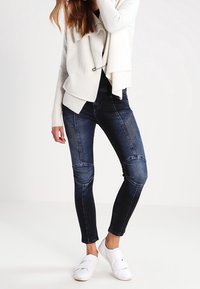 Mörkblå skinny jeans med knälappar, i kombination med en vit texturerad cardigan och vita slip-in skor.