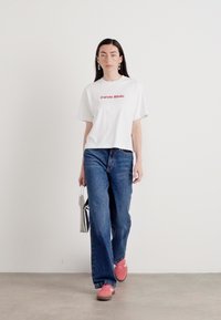 Baltas kokvilnas T-krekls ar sarkano "Calvin Klein" logotipu, kombinēts ar zilām denim platām biksēm un rozā kedām. Aksesuāri ietver svītrotu somu.