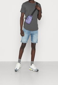 Lila nylon crossover-väska med justerbar rem, grå bomullströja, denimshorts och grå/vita sneakers med gula accenter.