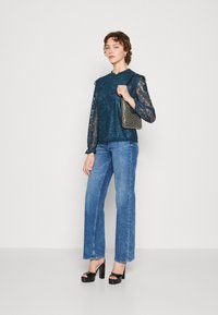 Bluza od čipke u tirkiznoj boji s dugim rukavima i valovitim rubom, uparena s plavim širokim trapericama i crnim platformskim štiklama. Dodatno je ukrašena malom torbicom.