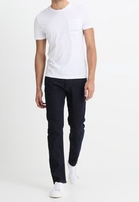 Vit bomullströja med bröstficka och logotyp, kombinerad med mörkblå jeans och vita sneakers, stående mot en ljus bakgrund.