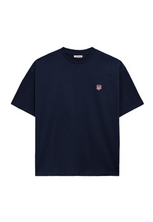 Junger Mann trägt eine cremefarbene und marineblaue Kappe und ein schwarzes T-Shirt mit kleinem Logo auf der linken Brust, kombiniert mit grauen Nadelstreifenhosen.