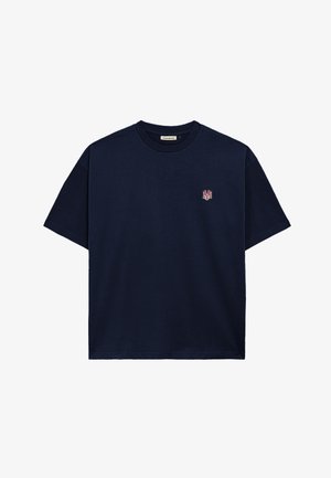 Jonge man die een crème- en marineblauwe pet draagt en een zwart T-shirt met een klein logo op de linkerborst, gecombineerd met grijze krijtstreepbroek.