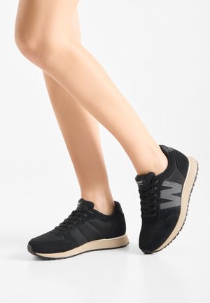 Persoon draagt zwarte mesh sneakers met beige zolen, één voet plat op de grond en de andere lichtjes op de tenen geheven.
