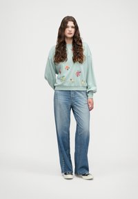 Világos zöld pulóver virágos hímzéssel, laza kék farmerral és fekete tornacipővel párosítva. Simogatós anyag, laza fazon, és sportos stílus.