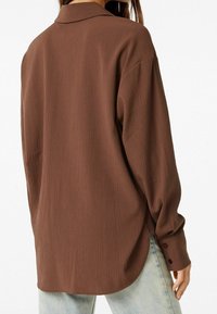 Camicia marrone a maniche lunghe in tessuto testurizzato, con una vestibilità rilassata, colletto e polsini con bottoni. Indossata con jeans blu chiaro.