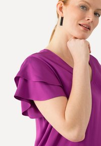 Lilla bluse med korte, lagdelte flæseærmer, V-udskæring og glat, let stof. Model poserer med hånden under hagen.