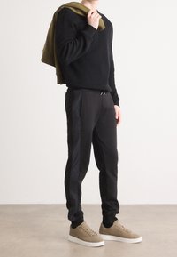 Suéter negro acanalado, joggers negros ajustados con rayas laterales y zapatillas de ante beige. Sudadera verde oliva colgada del brazo.