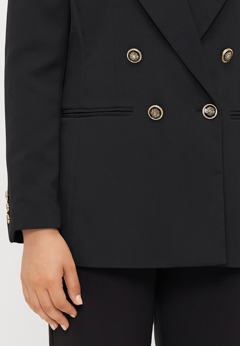 Schwarze doppelt geknöpfte Blazerjacke aus strukturiertem Stoff, mit goldenen runden Knöpfen, seitlichen Taschen und einem scharfen Kragen. Taillierte Passform mit strukturierten Schultern.