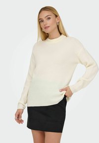 Cremfarbener gerippter Pullover mit hohem Kragen, langen Ärmeln und lockerer Passform, kombiniert mit einem schwarzen Rock. Glatte Textur.